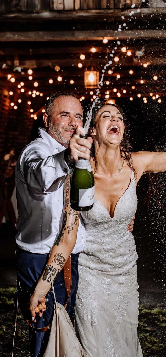 bride-and-groom-celebrating-champagne A bride and groom celebrate by spraying a bottle of champagne, smiling and laughing under string lights at an outdoor venue.