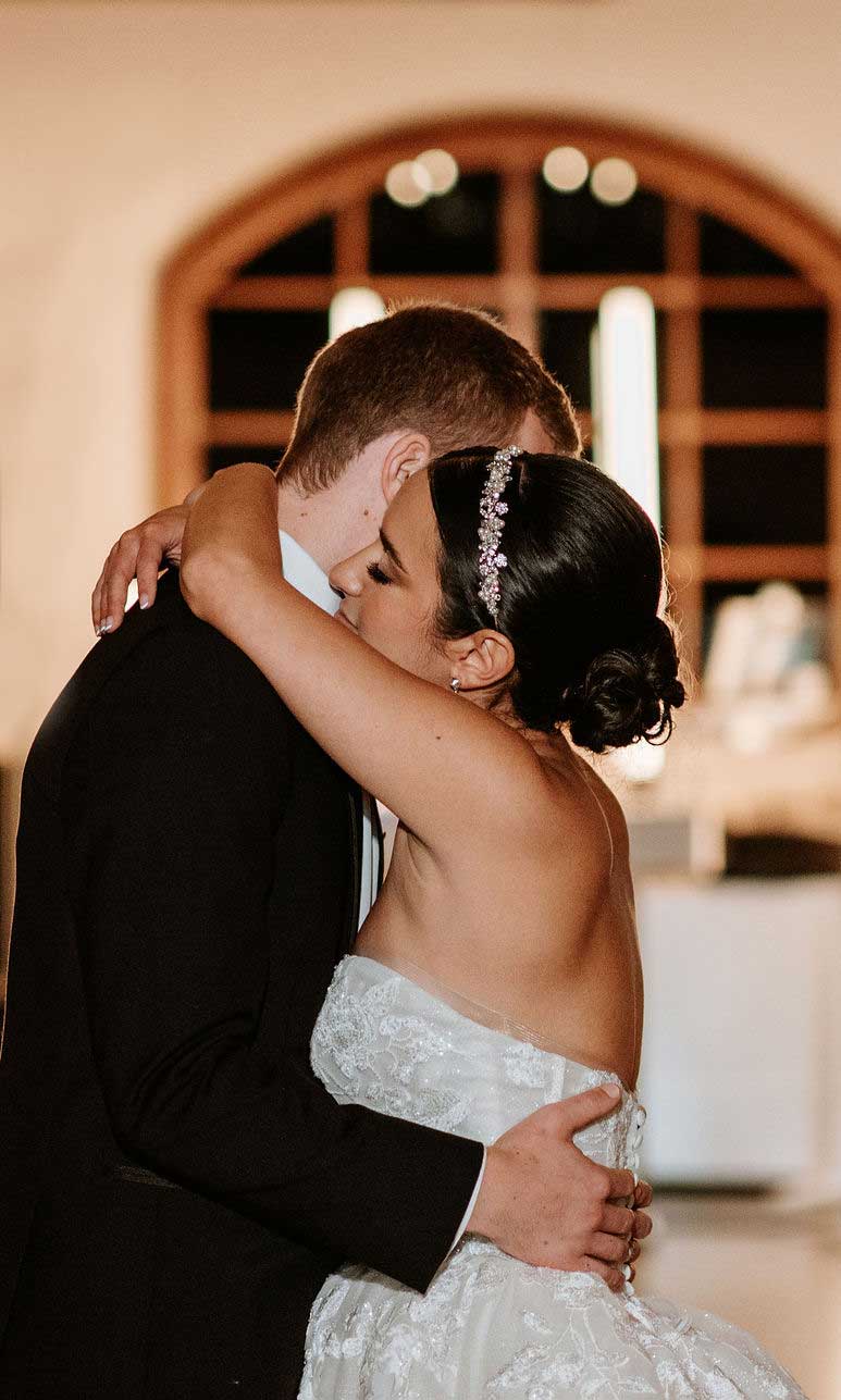 bride-and-groom-slow-dance A bride and groom embrace closely on the dance floor, both dressed in formal wedding attire, with a window and soft lighting in the background.