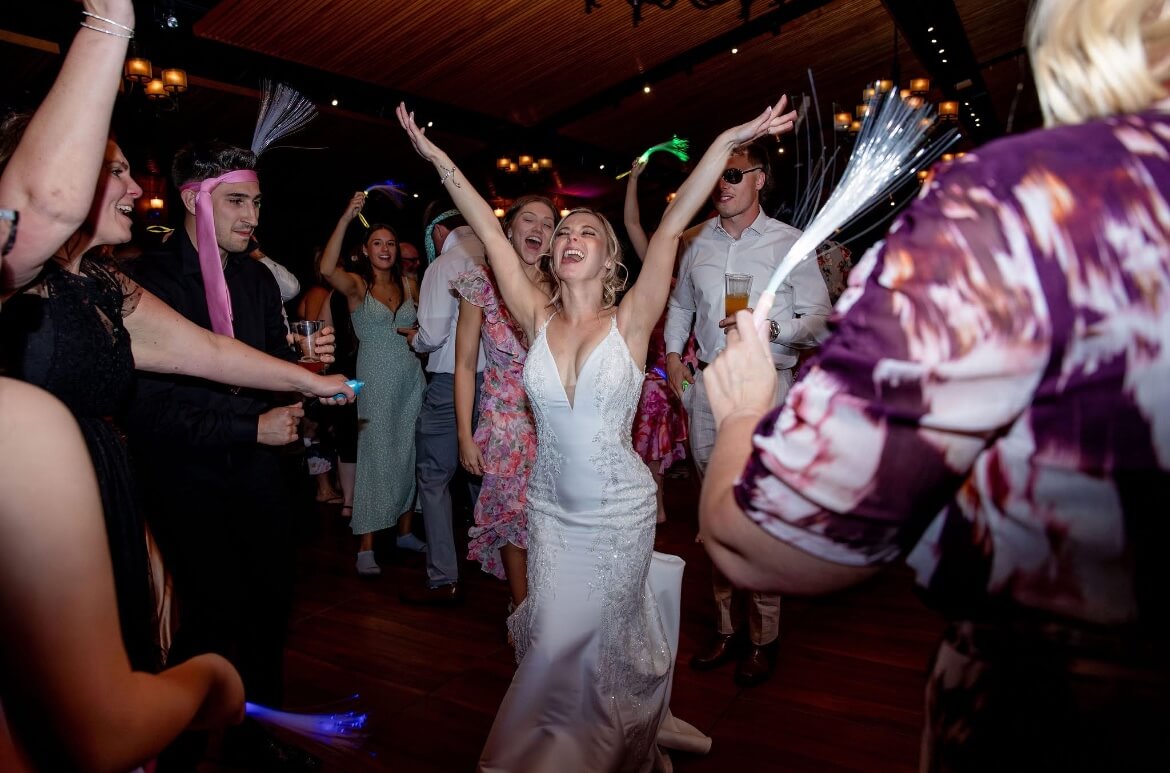 A bride in a white dress dances with her arms raised, surrounded by guests on a lively indoor dance floor.