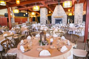 Elegant banquet hall with round tables set for an event, featuring folded napkins, floral centerpieces, and a stone fireplace in the background.