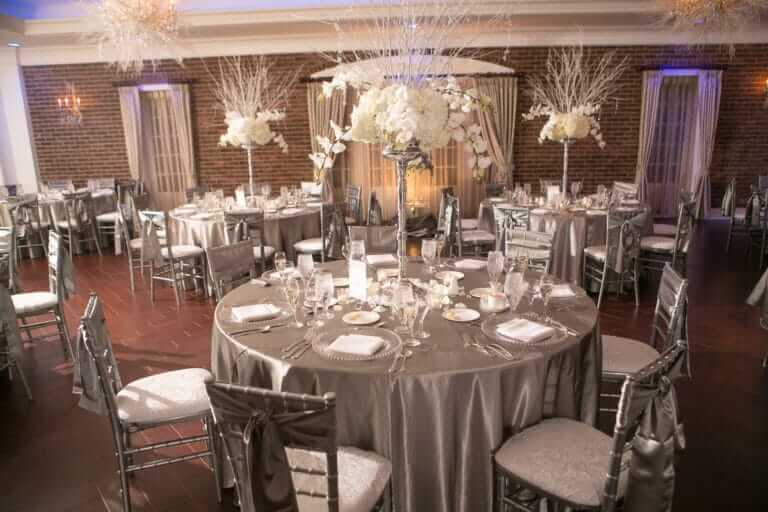 Elegant banquet hall with round tables covered in silver tablecloths, silver chairs, and white floral centerpieces, set for a formal event.