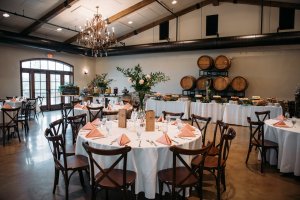 Elegant banquet hall set for an event, featuring round tables with white tablecloths, folded peach napkins, floral centerpieces, and a buffet with wine barrels in the background.