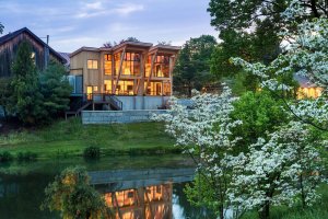 Modern two-story house with large windows sits beside a pond, surrounded by trees and blooming white flowers, at dusk.