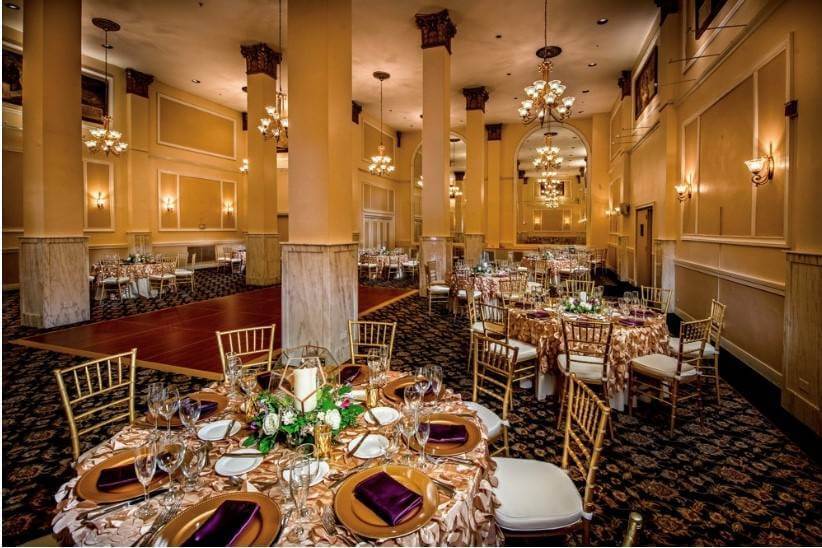 Elegant banquet hall with round tables set for an event, featuring gold chairs, purple napkins, floral centerpieces, and chandeliers overhead.
