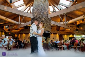 A bride and groom dance on a fog-covered floor in a rustic, wooden venue with guests seated around them.