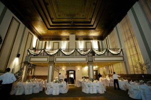 A large, elegant banquet hall with high ceilings, decorated tables and chairs, draped white fabric along the balcony, and a few people setting up for an event.