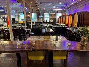 A spacious event venue with wooden tables, chairs, floral centerpieces, string lights, large wine barrels, and people setting up in the background.