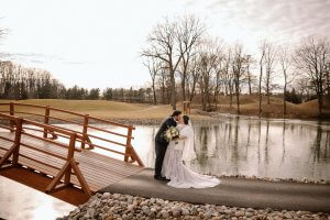 A bride and groom stand by a wooden bridge next to a pond, sharing a kiss on a cloudy day with bare trees in the background.