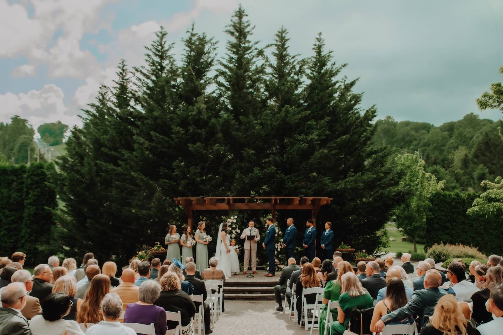 Bear-Creek-22 An outdoor wedding ceremony with a bride, groom, officiant, and wedding party standing under a wooden arbor, surrounded by seated guests and tall evergreen trees.