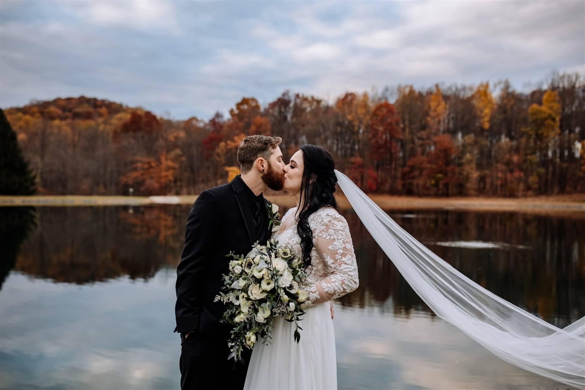 Nicole + Justin // Wedding Day A bride and groom kiss by a lake with autumn trees in the background; the bride holds a bouquet and her veil flows in the breeze.