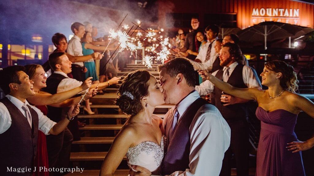 A bride and groom kiss on a staircase while guests surround them holding sparklers, celebrating at a nighttime wedding reception.